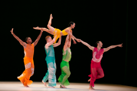 4 dancers in brightly colored costumes lift another dancer above their heads.